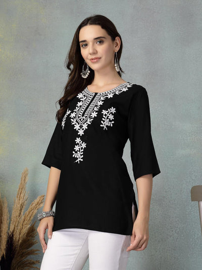 Woman wearing black embroidered rayon kurti with white floral designs paired with white pants against a grey backdrop