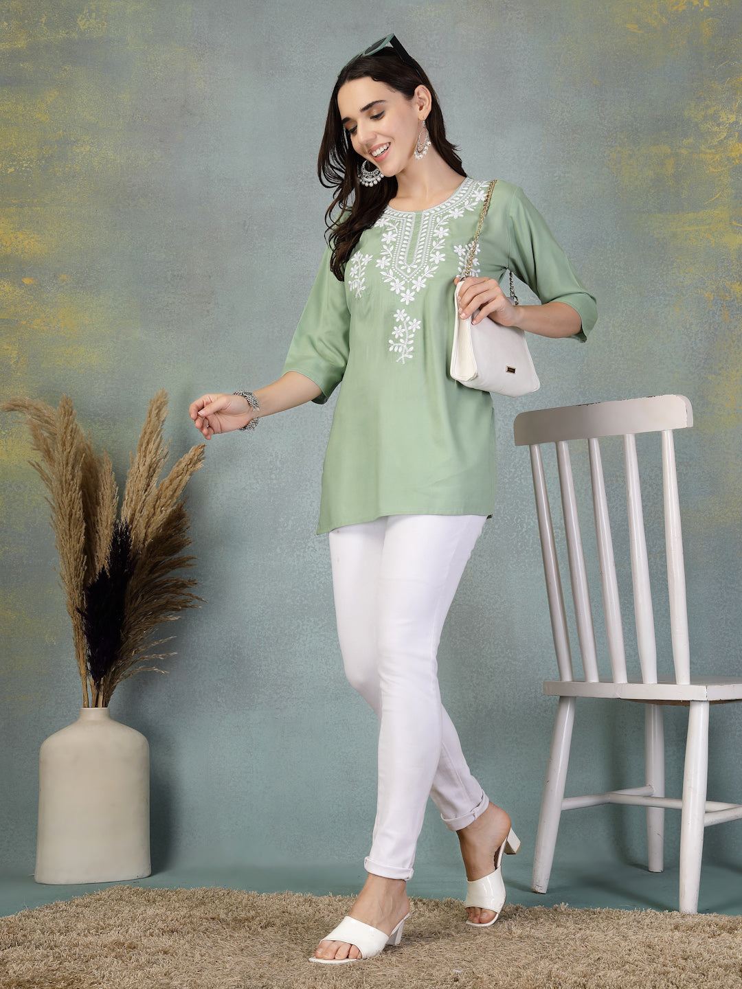 Woman wearing pista green embroidered rayon kurti paired with white leggings and white sandals, standing near a chair and vase with dried plants.