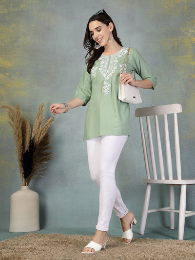 Woman wearing pista green embroidered rayon kurti paired with white leggings and white sandals, standing near a chair and vase with dried plants.